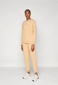 Svetlo bež pulover in usklajene trenirka iz mehke tkanine. Ima sprednji žep, rebraste manšete in majhen logotip na prsih.