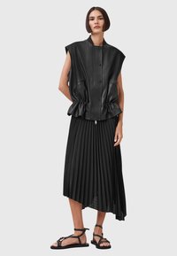 Black leather sleeveless jacket with front zip, pleated black skirt, and black strappy sandals. Features a cinched waist and multiple pockets.