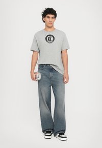 Tricou din bumbac gri cu imprimeu grafic negru, asortat cu blugi largi de culoare albastru deschis. Modelul ține un accesoriu argintiu. Sneakerși alb-negru.