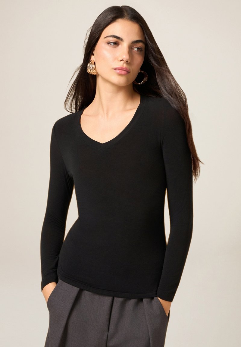 Czarna bluzka z długim rękawem i dekoltem w serek, wykonana z gładkiego materiału, o dopasowanym kroju i subtelnym połysku. Połączona z wysokim stanem szarymi spodniami.