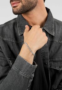 Bracciale d'argento con molte pietre preziose trasparenti, indossato al polso. La persona indossa una giacca di denim scuro con collo abbottonato.