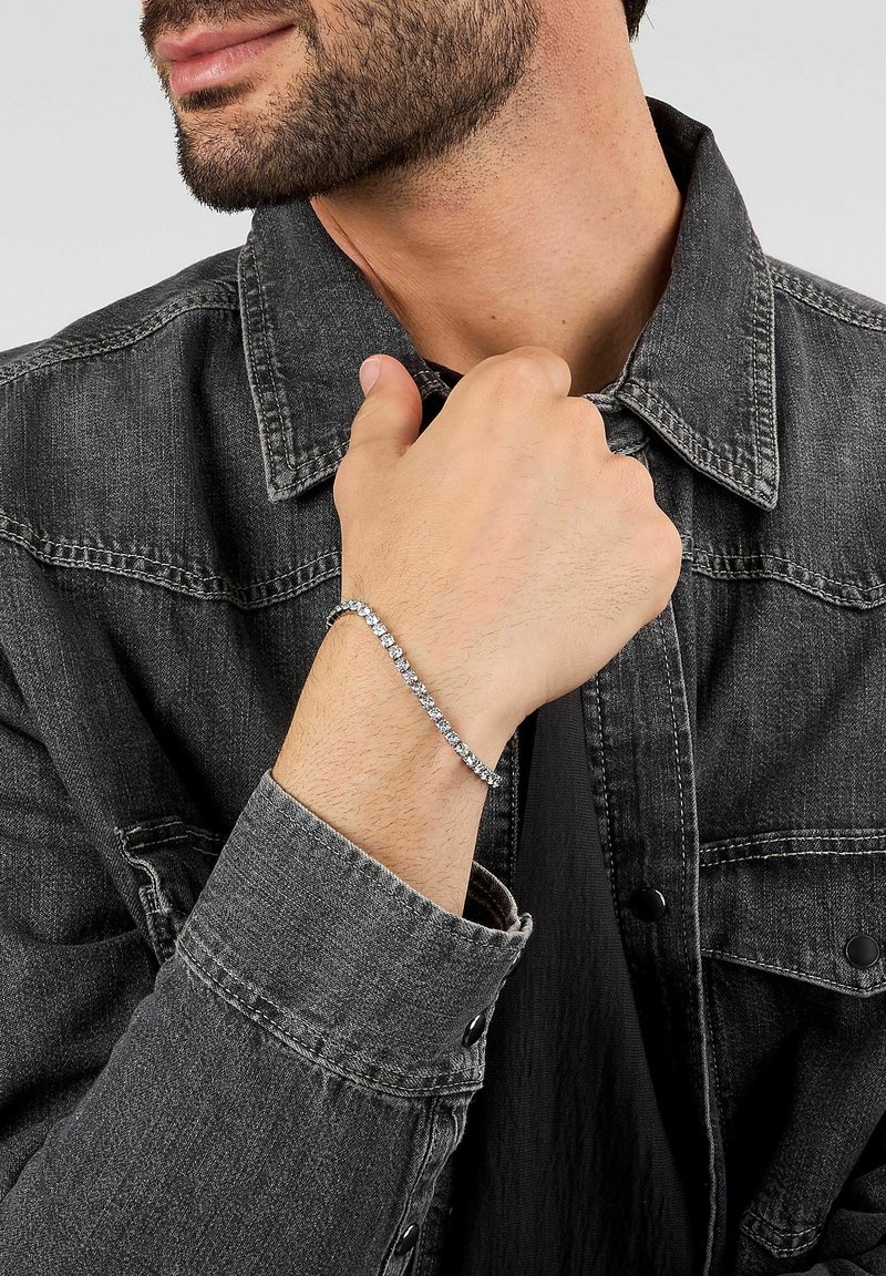 Bracciale d'argento con molte pietre preziose trasparenti, indossato al polso. La persona indossa una giacca di denim scuro con collo abbottonato.