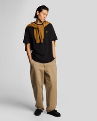 Melnā T-krekls ar nelielu logotipu, bēša plata slēguma bikses un brūns džemperis pār pleciem, kopā ar melniem apaviem.