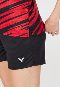 Sorte træningsshorts med et gråt logo og en rød top med zigzagmønster langs siden, lavet af et glat, let stof.