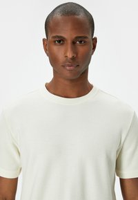 Ljuskrämfärgad t-shirt med korta ärmar i en texturerad, nätliknande tyg. Har en rund halsringning och minimala designelement.