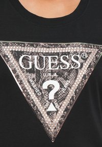 Maglietta nera con un luccicante logo triangolare GUESS che presenta un motivo a serpente, bordi borchiati e il testo "U.S.A. Washed Jeans" con un punto interrogativo.