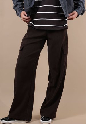 Personne portant un pantalon cargo noir à jambes larges, une chemise rayée noire et blanche, une veste sombre et des baskets noires avec des semelles blanches.