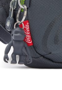 Borsa in tessuto nero con un portachiavi in plastica grigia a trama liscia raffigurante una figura con le braccia tese. È visibile una targhetta rossa Coca-Cola.