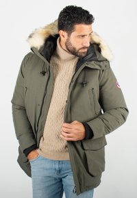 Parka olive verte avec capuche en fourrure, accessoires noirs, fermeture éclair et poignets côtelés. Porté par-dessus un pull en laine torsadée crème.