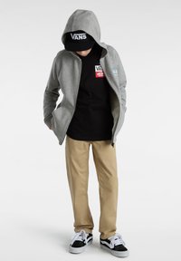 Sudadera gris con cremallera y capucha, camiseta negra con logo, pantalones beige y zapatillas negras y blancas. Presenta líneas simples y un estilo casual.