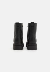 Une paire de bottines en cuir noir, vues de l'arrière, dotées de semelles épaisses et de fermetures éclair visibles sur un fond blanc.