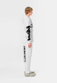 Sudadera blanca de gran tamaño con gráficos en negro, combinada con pantalones blancos que presentan impresiones laterales y zapatos texturizados en gris.