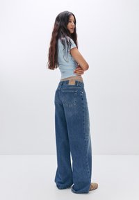 Breda denimjeans med en ljusblå bomullströja. Har en ljusbrun lappar på baksidan och en blekt blå tvätt.