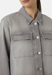 Graue Jeansjacke mit zwei Brusttaschen, Knopfverschluss und einem umgelegten Kragen. Der Stoff hat eine glatte Textur mit einem leicht verblassten Finish.