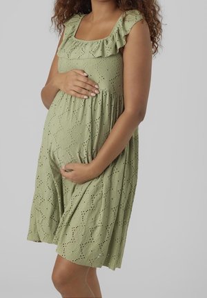 Schwangere Frau in einem hellgrünen Lochkleid, die ihren Bauch sanft mit beiden Händen vor einem schlichten grauen Hintergrund hält.