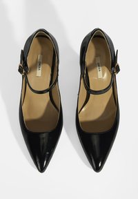 Schwarze Mary Jane Pumps aus Lackleder mit spitzen Zehen und verstellbarem Knöchelriemen mit goldener Schnalle. Innenraum ist beige.