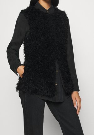 Vrouw draagt een mouwloos zwart zacht vest over een geknoopt zwart overhemd en zwarte broek, één hand in het vestzakje.