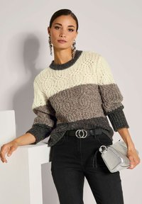 Frau in einem strukturierten, mehrfarbigen Pullover in Creme, Taupe und Anthrazit, schwarzen Hosen mit einem Doppelring-Gürtel, silbernen Ohrringen und einer silbernen Handtasche.