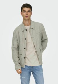 Hellgraue Button-up-Jacke mit strukturiertem Stoff, ausgestattet mit zwei Fronttaschen und einem lockeren Schnitt, getragen über einem hellbeigen Hemd und blauen Jeans.