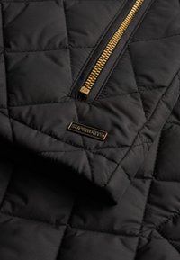 Czarna pikowana kurtka z złotym zamkiem i metalowym logo Superdry. Tkanina ma gładką strukturę z pikowaniem w kształcie diamentów.