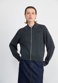 Grijze kabelgebreide cardigan met een rits aan de voorkant en een kraaghals. Heeft ribgebreide manchetten en zoom, gecombineerd met een donkere denim rok.