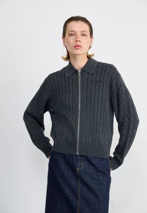 Gray cable-knit cardigan with a zip front and collared neckline. Features ribbed cuffs and hem, paired with a dark denim skirt.
