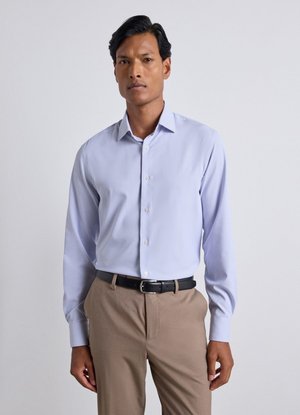 Mann trägt ein hellblaues Hemd, das in beige Hose mit schwarzem Gürtel gesteckt ist, und steht vor einem einfarbigen hellen Hintergrund.