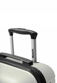 Hard-shell suitcase in light cream with a textured surface, black zipper, and a telescopic handle with a rubber grip. "Bench." logo visible.