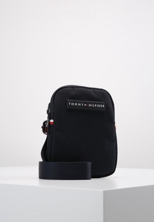 Kleine zwarte Tommy Hilfiger crossbodytas met verstelbare riem en logopatch aan de voorkant, geplaatst op een witte ondergrond.