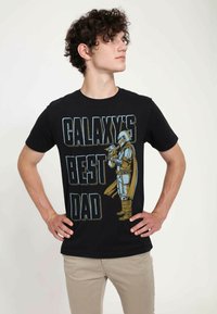 Černé bavlněné tričko s výrazným textem "NEJLEPŠÍ OTEC GALAXIE" a barevným ilustrovaným charakterem v brnění, který drží malou postavičku.