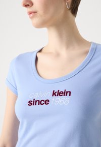 Light blue short-sleeve t-shirt with a scoop neck and embroidered text: "calvin klein" in white and "since 1968" in burgundy. Soft fabric.