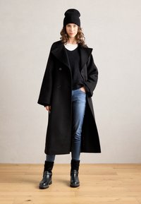 Abrigo largo de lana negra con amplios solapas, botones de doble botonadura, llevado sobre un suéter negro y jeans claros. Gorro de lana negro y botas de tobillo.