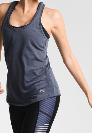 Frau trägt ein ärmelloses, dunkelgraues Sport-Top und schwarze Leggings mit blauen Streifendetails, posiert mit einem ausgestreckten Arm.