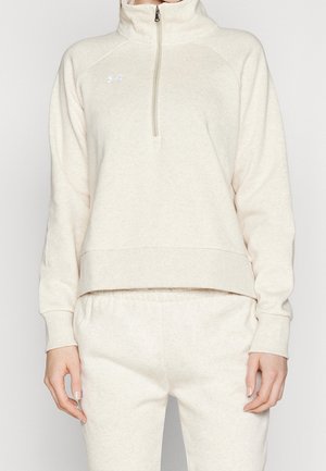 Vrouw draagt een lichtbeige sweater met kwartritssluiting en bijpassende joggingbroek, staand tegen een effen achtergrond.