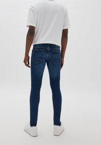 Man i mörkblå smala jeans, en enkel vit t-shirt och vita sneakers, stående med ryggen mot en neutral bakgrund.