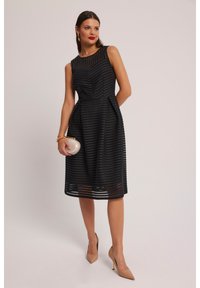 Czarna sukienka na ramiączkach bez rękawów, z żebrowaną teksturą, dopasowanym gorsetem, rozkloszowanym dołem oraz przezroczystymi akcentami. Modelka trzyma okrąłą srebrną kopertówkę i ma na sobie cieliste szpilki.