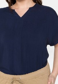 Námořnická modrá polokošile s krátkým rukávem a výstřihem do V, zdobená jemným vertikálním švem, v kombinaci s béžovými šortkami na stahování. Měkká, lehká tkanina.