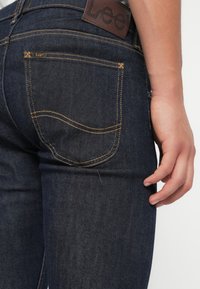 Jeans de mezclilla oscura con un ajuste ceñido, costuras naranjas y una etiqueta de cuero marrón en la parte trasera. Cuenta con dos bolsillos traseros con bordes sutilmente curvados.