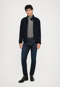 Navy zip jacket over a gray polo, paired with dark blue jeans and black shoes. Smooth fabric, modern fit with a collar detail.