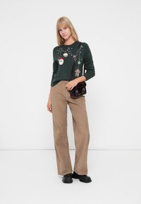 Vihreä neulottu villapaita, jossa on jouluteemaisia koristeita, yhdistetty vaaleanbeigeihin samettihousuihin, joissa on leveät lahkeet, sekä pieneen tummaan käsilaukkuun. Mustat kengät.