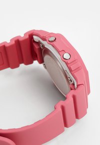 Bracelet de montre en silicone rose avec une finition texturée, présentant des boutons en argent et un revêtement intérieur blanc, révélant partiellement le boîtier arrière en métal.
