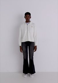 White zip-up hoodie with drawstrings, paired with dark flared jeans. Model stands in neutral background, wearing white sneakers.