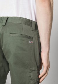 Zelené cargo kalhoty mají zadní kapsu a malou značkovou nášivku v červené, bílé a modré barvě na horní zadní straně. Hladká struktura látky.