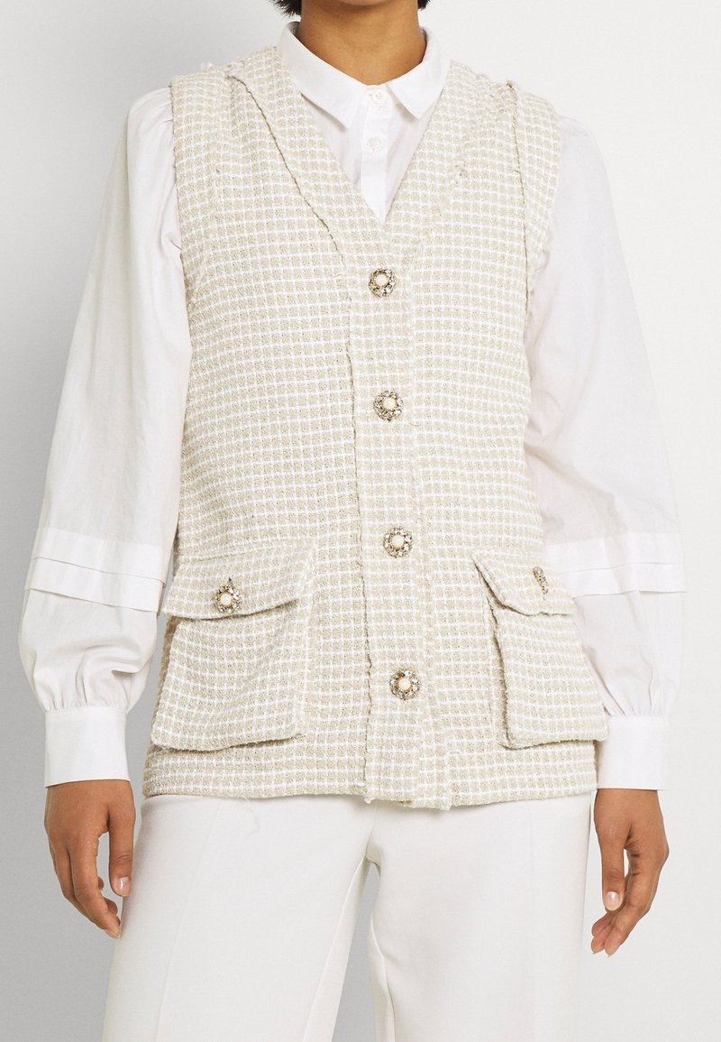 Femme portant un gilet sans manches texturé crème avec des boutons en perle sur un chemisier blanc à manches longues et un pantalon blanc.
