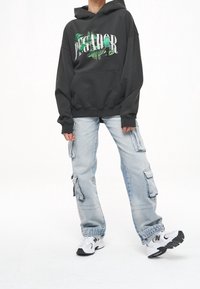 Sudadera negra con estampado gráfico verde y bolsillo frontal, combinada con jeans cargo azul claro que presentan múltiples bolsillos y un corte holgado. Zapatillas blancas.