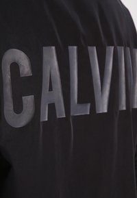 Czarna tekstylna kurtka z dużymi, szarymi, gumowanymi literami napisanymi "CALVIN" na plecach. Gładka struktura, sportowy krój i minimalistyczny styl.