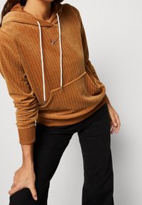 Brun kordflod hoodie med vita dragsko, ribbade ärmar, framficka och subtil logodetalj på bröstet; textilstruktur och avslappnad passform.