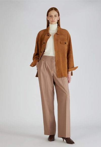 Veste en daim marron avec des boutons, col roulé blanc, pantalon plissé beige et chaussures à talons marron sur un fond uni.