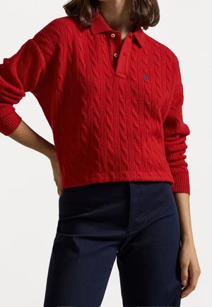 Femme portant un pull polo rouge en maille torsadée à manches longues avec un pantalon bleu foncé, main sur la hanche, sur un fond clair uni.