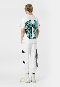 Camiseta blanca de manga corta con grandes gráficos de alas en negro y turquesa en la parte trasera, combinada con joggers blancos y botas blancas gruesas.